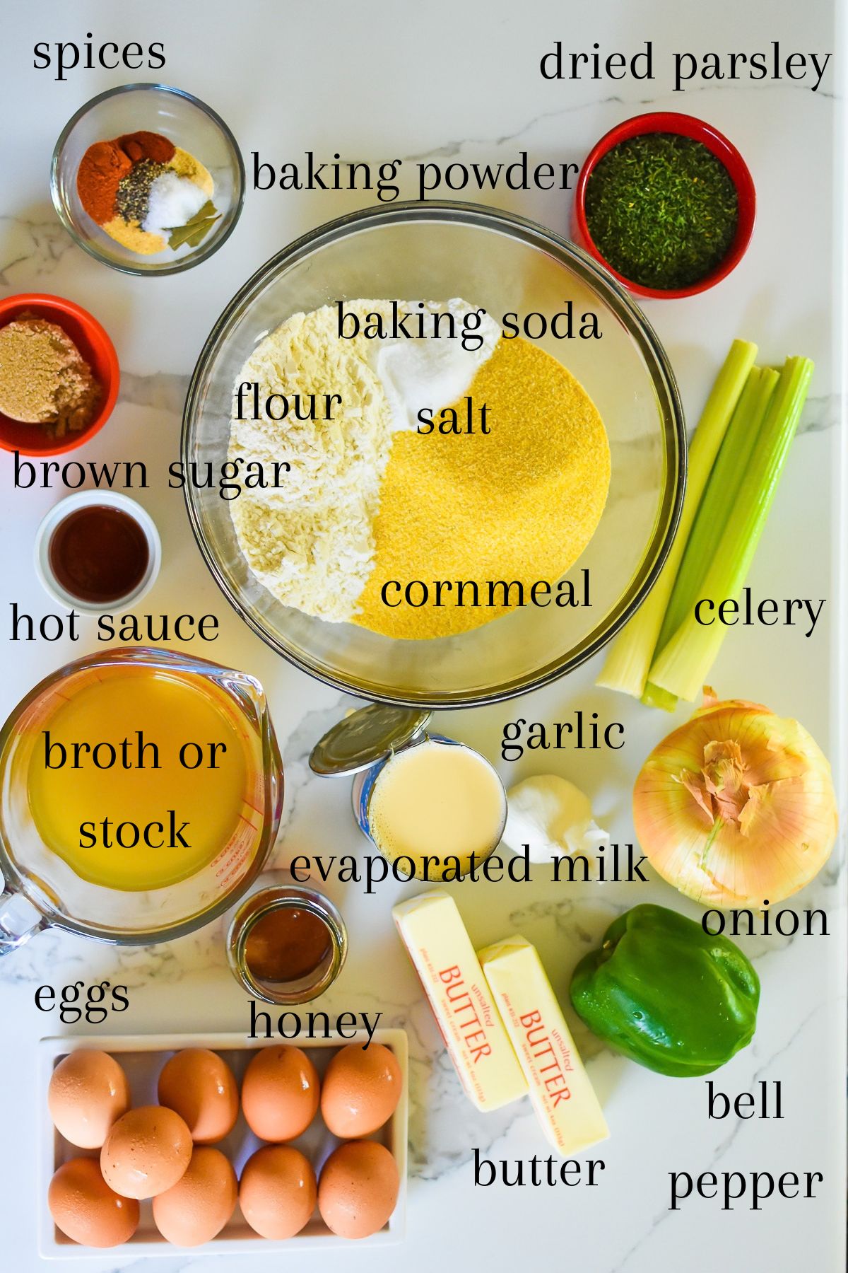 ingredients for making a cornbread dressing recipe Soul Food style on granite counter top: spices, brown sugar, dried parsley, cornmeal, flour, baking powder, baking soda, salt, celery, onion, garlic, evaporated milk, hot sauce, chicken broth, bell pepper, honey, unsalted butter, and eggs.