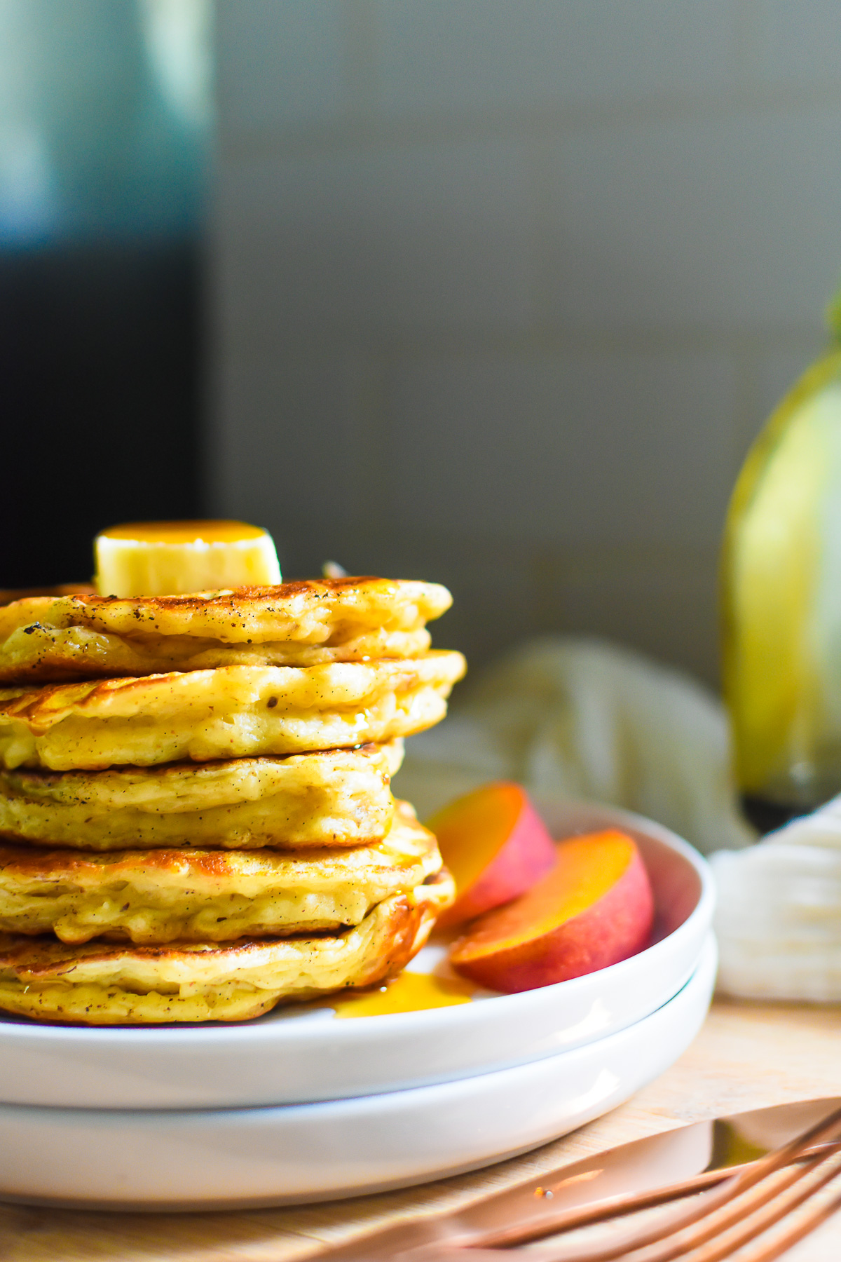 five Greek yogurt thick pancakes stacked on a plate, topped with maple syrup and a pat of butter, with peach slices on the side.