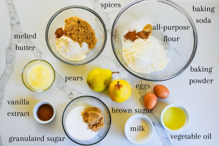 ingredients for making a moist pear cake recipe from scratch on kitchen counter top: flour, spices, baking soda and powder, brown sugar, granulated sugar, eggs, vegetable oil, milk, fresh pears, melted butter, and granulated sugar.